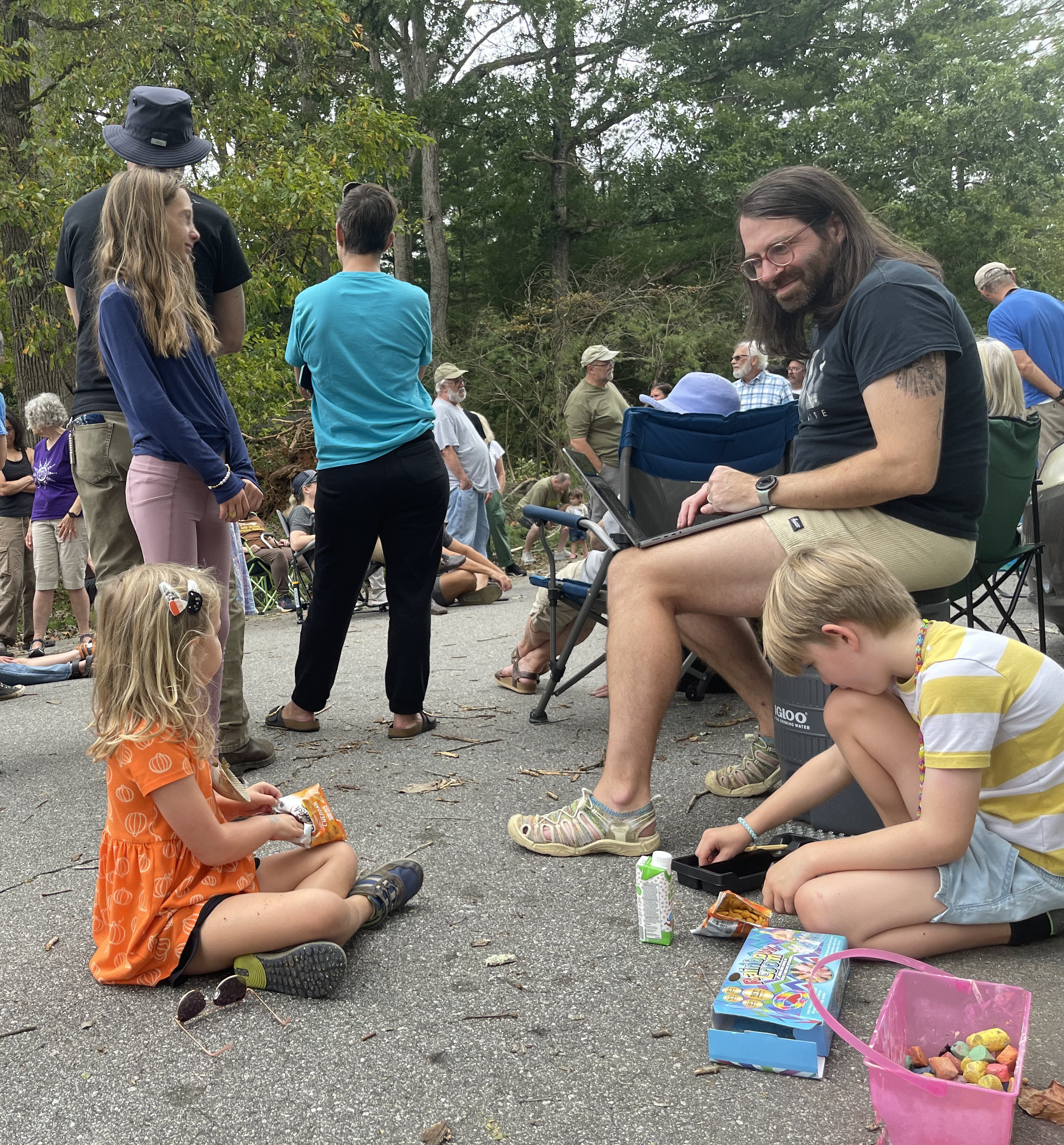 Dan Ferrell, a white person with long hair and glasses, wearing a black shirt and shorts, typing notes on a computer during a post-hurricane organizing meeting.